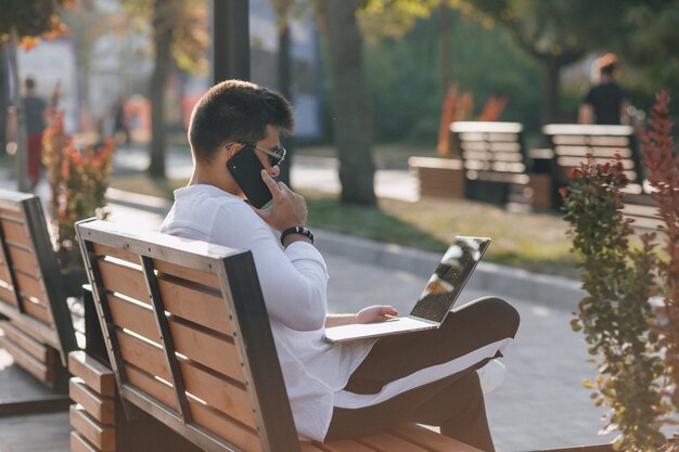 Młody stylowy facet w koszuli z telefonu i notebooka na ławce w słoneczny dzień na zewnątrz