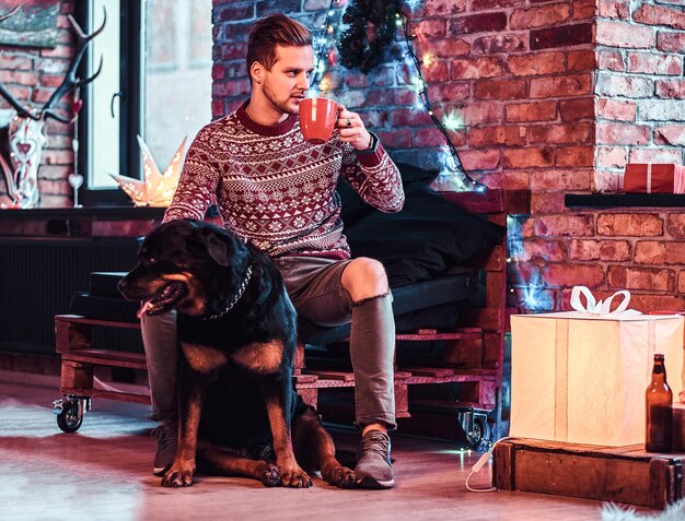 Bezpłatne zdjęcie młody stylowy mężczyzna trzyma kubek z kawą w czasie świąt bożego narodzenia ze swoim uroczym psem w urządzonym salonie.