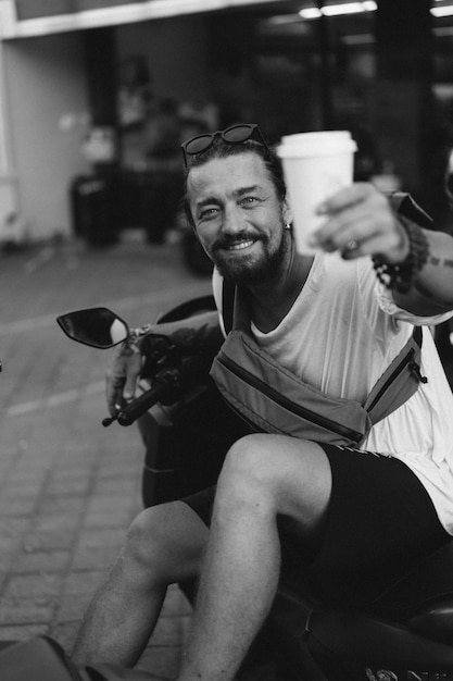 Młody stylowy mężczyzna z brodą pije kawę z jednorazowego papierowego kubka, siada na motocyklu na ulicy. Styl życia, życie tropikalne. Bali