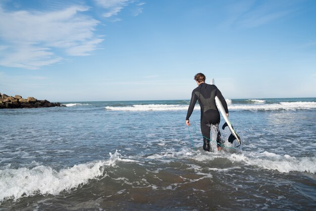 Młody surfer wchodzący do wody z deską surfingową w czarnym kombinezonie. Koncepcja sportu i sportów wodnych.