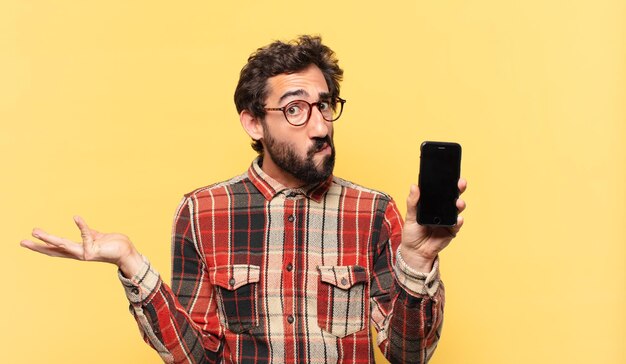 Młody szalony brodaty mężczyzna wątpiący lub niepewny wyraz twarzy i trzymający telefon