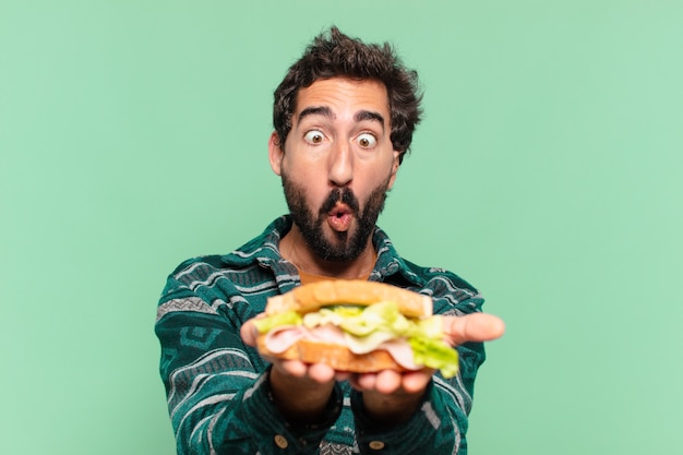 Młody szalony brodaty mężczyzna ze szczęśliwym wyrazem twarzy i trzymający kanapkę