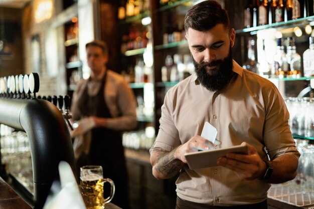Młody szczęśliwy barista pracujący w barze i surfujący po sieci na touchpadzie.