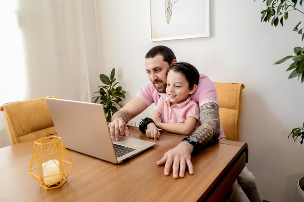 Młody szczęśliwy ojciec w różowej koszulce polo pracuje na laptopie siedząc przy stole ze swoim synkiem szczęśliwa rodzina pracuje w domu koncepcja