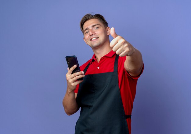 młody uśmiechnięty blond mężczyzna fryzjer w mundurze posiada telefon kciuki do góry na białym tle na fioletowym tle