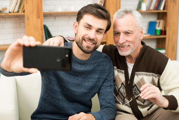 Młody uśmiechnięty facet i starzejący się rozochocony mężczyzna bierze selfie na smartphone na kozetce