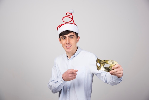 Młody uśmiechnięty mężczyzna model stojący w świątecznym kapeluszu wskazujący na karnawałową maskę