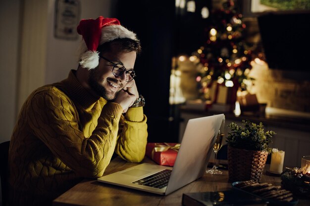 Młody uśmiechnięty mężczyzna za pomocą laptopa w Wigilię Bożego Narodzenia w domu