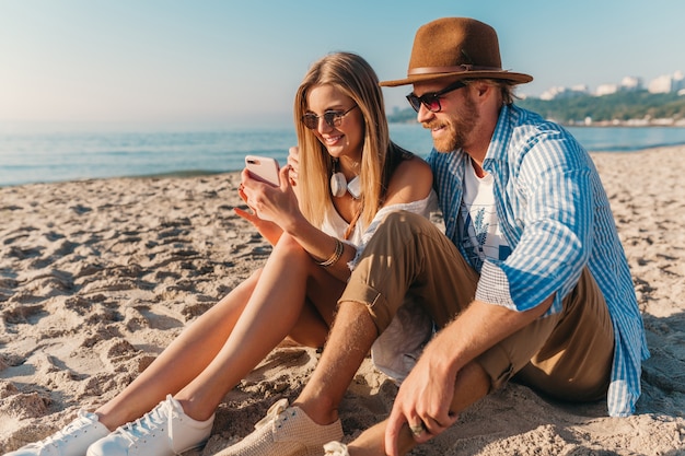 Młody uśmiechnięty szczęśliwy mężczyzna i kobieta w okularach przeciwsłonecznych siedzi na piaszczystej plaży, biorąc zdjęcie selfie na aparat w telefonie