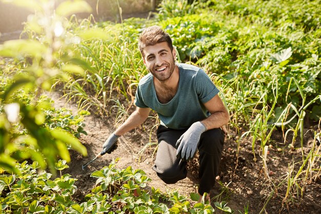 Młody wesoły atrakcyjny, brodaty męski ogrodnik w niebieskiej koszulce i czarnych spodniach, uśmiechnięty, pracujący w ogrodzie, sadząc kiełki z łopatą.