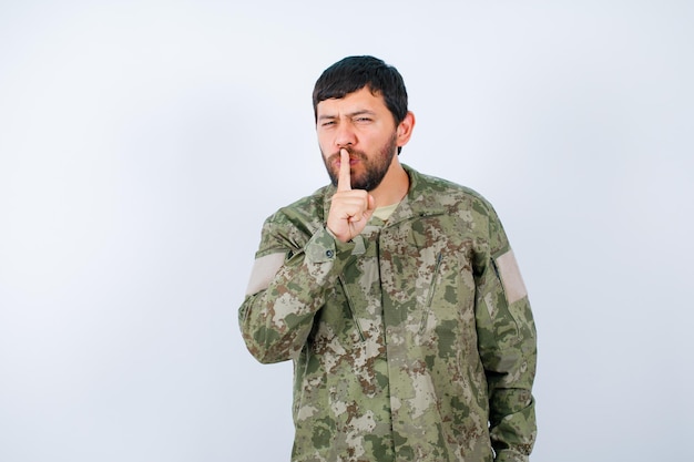 Młody wojskowy pokazuje gest ciszy, trzymając palec wskazujący na ustach na białym tle