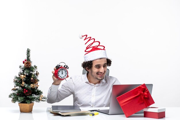 Młody zabawny emocjonalny podekscytowany biznesmen z Santa Claus kapelusz i trzymając i pokazując zegar siedzi w biurze na białym tle stock photo