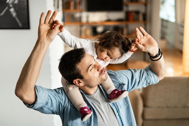 Młody, zabawny ojciec bawi się, niosąc w domu swoją małą córeczkę na ramionach