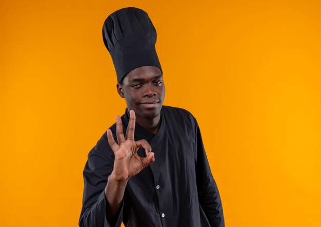 Młody zadowolony afro-amerykański kucharz w mundurze szefa kuchni gestów ok ręka znak na białym tle na pomarańczowym tle z miejsca na kopię