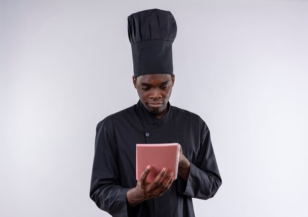 Młody zadowolony afro-amerykański kucharz w mundurze szefa kuchni trzyma i patrzy na notebook na białym z miejsca na kopię