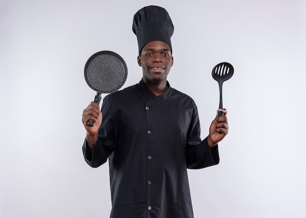 Młody zadowolony afro-amerykański kucharz w mundurze szefa kuchni trzyma patelnię i łopatkę na białym tle z miejsca na kopię