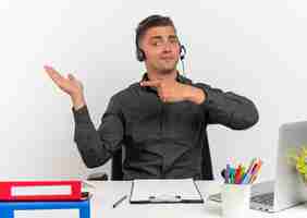 Bezpłatne zdjęcie młody zadowolony blond pracownik biurowy mężczyzna na słuchawkach siedzi przy biurku z narzędzi biurowych za pomocą punktów laptopa w pustej dłoni
