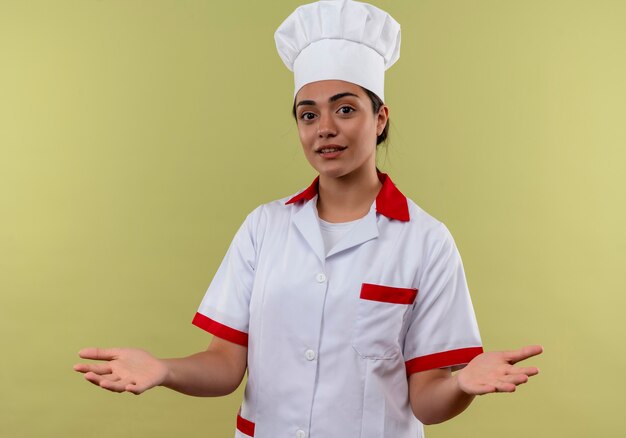 Młody zadowolony kucharz kaukaski dziewczyna w mundurze szefa kuchni trzyma ręce otwarte na białym tle na zielonej ścianie z miejsca na kopię