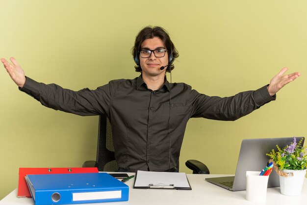 młody zadowolony pracownik biurowy mężczyzna na słuchawkach w okularach siedzi przy biurku za pomocą laptopa trzyma otwarte ramiona
