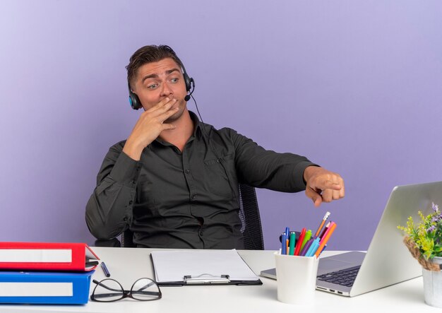Młody zaskoczony blond pracownik biurowy mężczyzna na słuchawkach siedzi przy biurku z narzędziami biurowymi patrząc i wskazując na laptopa kładzie dłoń na ustach na fioletowym tle z miejsca na kopię