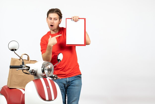 Młody zaskoczony facet dostawy w czerwonym mundurze, stojący w pobliżu skutera, wskazując dokument na białej ścianie