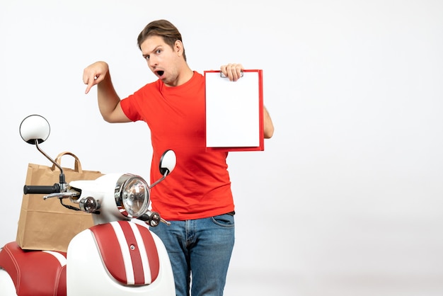 Bezpłatne zdjęcie młody zaskoczony facet dostawy w czerwonym mundurze stojącym w pobliżu skutera przedstawiający dokument wskazujący papierową torbę na białej ścianie