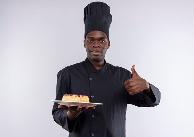 Młody zaskoczony kucharz afro-amerykański w mundurze szefa kuchni trzyma ciasto na talerzu i kciuki do góry na białym z miejsca na kopię