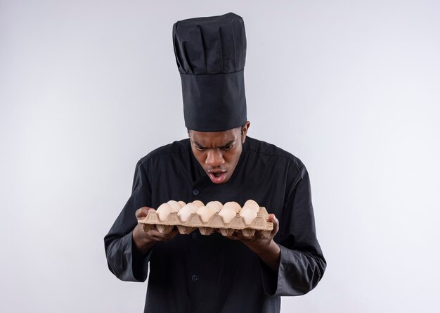 Bezpłatne zdjęcie młody zaskoczony kucharz afro-amerykański w mundurze szefa kuchni trzyma partię jaj na białym tle na białej ścianie