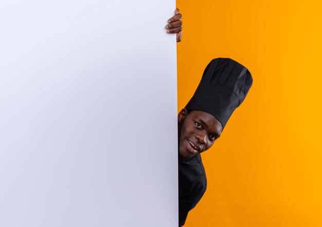 Młody zaskoczony kucharz afroamerykański w mundurze szefa kuchni stoi za białą ścianą odizolowaną na pomarańczowej przestrzeni z miejsca na kopię