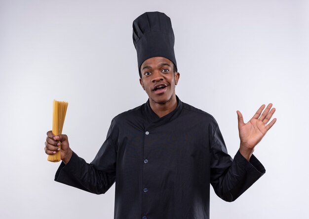 Młody zaskoczony kucharz afroamerykański w mundurze szefa kuchni trzyma kilka spaghetti i trzyma rękę na białym tle na białej ścianie