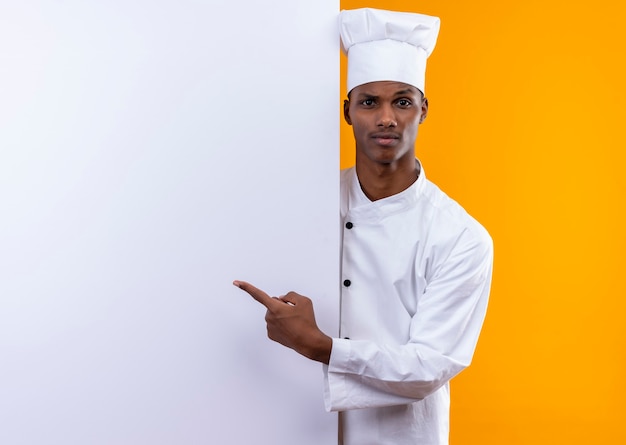 Młody Zdezorientowany Afro-amerykański Kucharz W Mundurze Szefa Kuchni Stoi Za Białą ścianą I Wskazuje Na ścianę Odizolowaną Na Pomarańczowej ścianie