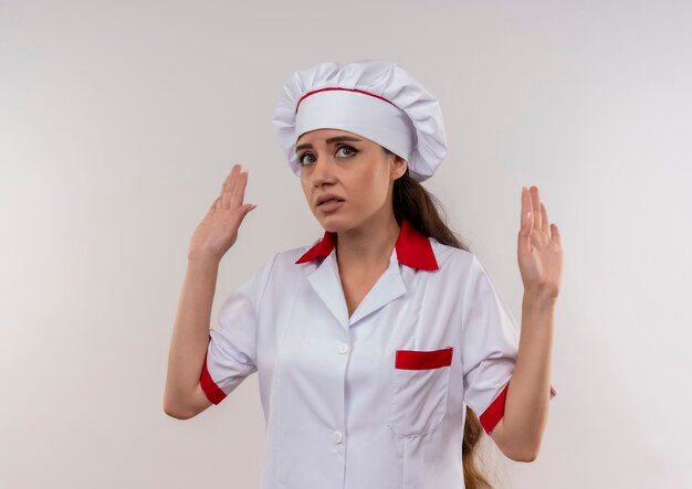 Młody zirytowany kucharz kaukaski dziewczyna w mundurze szefa kuchni podnosi ręce na białym tle na białym tle z miejsca na kopię