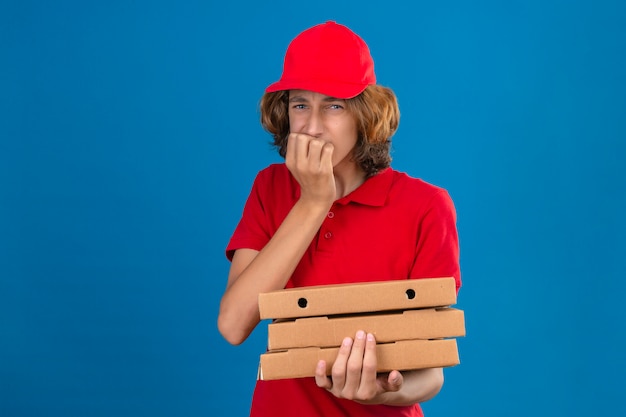 Młody zmartwiony dostawca w czerwonym mundurze trzymający pudełka po pizzy wyglądający na zestresowanego i zdenerwowanego, z rękami na ustach gryzącymi paznokcie na izolowanym niebieskim tle