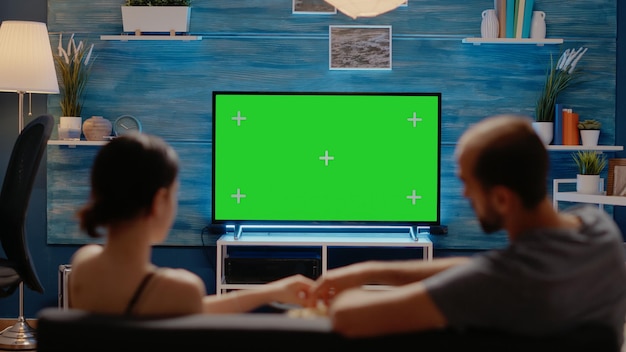 Młodzi ludzie oglądają telewizję z zielonym ekranem, siedząc w salonie. Kaukaski mężczyzna i kobieta z kluczem chromatycznym lub miejscem na kopię dla izolowanego szablonu i szablonu makiety