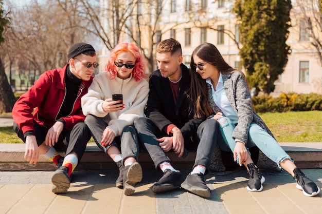 Młodzi ludzie siedzi na krawężniku i patrzeje smartphone