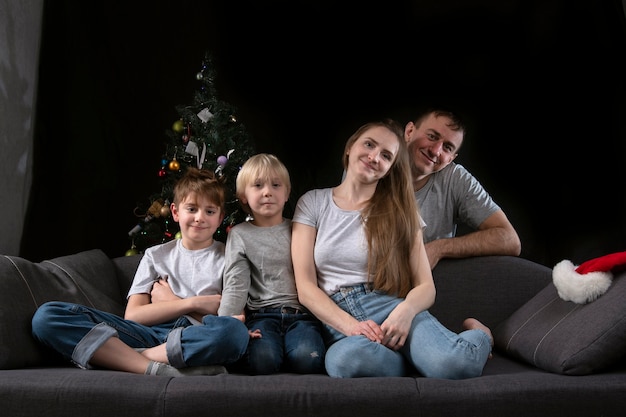 Młodzi rodzice i dwaj synowie siedzą w ciemnym salonie na kanapie w pobliżu choinki w wigilię bożego narodzenia.