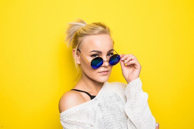 Moda blondynka w nowoczesny biały sweter w genialnych niebieskich okularach przeciwsłonecznych pozuje na żółto
