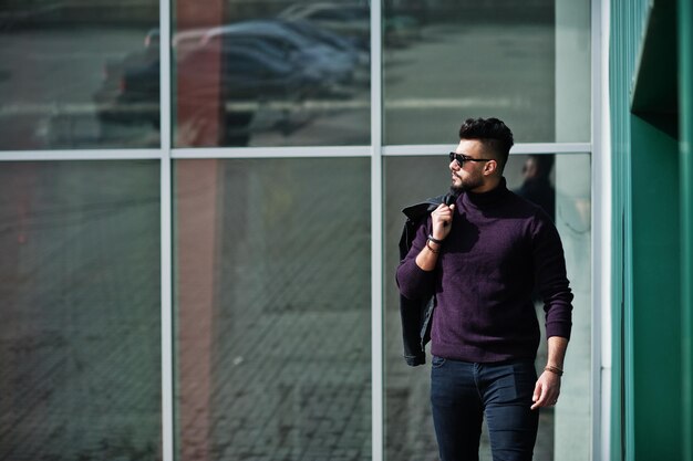 Moda bogata broda Arabski mężczyzna nosi golf i okulary przeciwsłoneczne postawione przed dużymi oknami nowoczesnego budynku z kurtką pod ręką Stylowy, udany i modny arabski model faceta