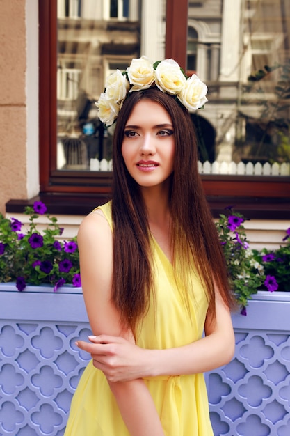 Bezpłatne zdjęcie moda portret szczęśliwy figlarny ładna brunetka dziewczyna uśmiecha się i dobrze się bawi, ubrana w pastelową żółtą sukienkę i wieniec róż.