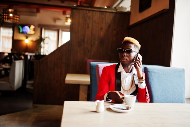 Model african american man w czerwonym garniturze z pasemkami włosów i okularami przeciwsłonecznymi, siedząc w kawiarni z kawą i rozmawiając przez telefon komórkowy