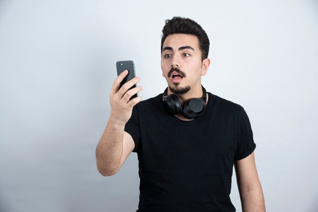 Model brunetka mężczyzna stojący w słuchawkach i przy użyciu telefonu na białej ścianie.