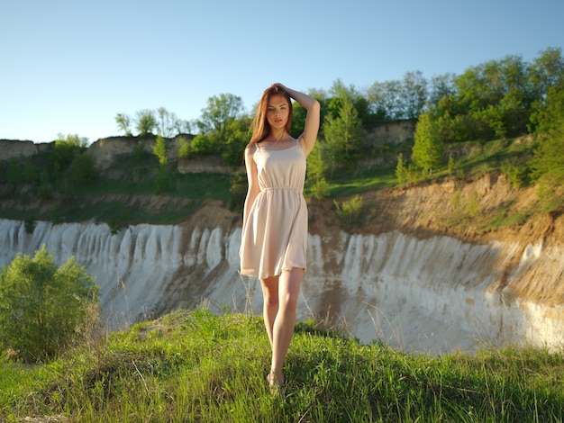 Bezpłatne zdjęcie modelka pozuje w słoneczny dzień z pięknym słonecznym krajobrazem wokół niej. młoda kobieta stojąca przy klifie z ładnym widokiem za plecami. atrakcyjna dziewczyna w białej sukni, pozowanie na zewnątrz.