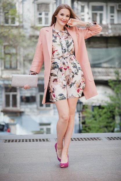 Modna atrakcyjna stylowa uśmiechnięta kobieta spaceru ulicą miasta w różowym płaszczu wiosenny trend w modzie trzymając torebkę