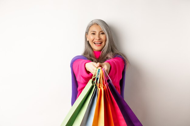Modna azjatycka starsza kobieta idzie na zakupy, wyciągając ręce z papierowymi torebkami, uśmiechając się zadowolona do kamery, stojąc na białym tle.