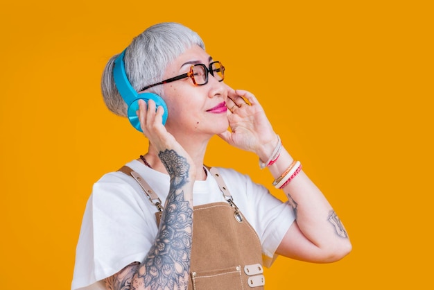 Modna azjatycka starsza kobieta nosić słuchawki słuchające melodii muzyki podczas noszenia fartuch ręka dotykać słuchawki dorywczo zrelaksować się styl życiałatwe słuchanie muzyki azja kobieta z tatuażem blisko oko czuć się swobodnie