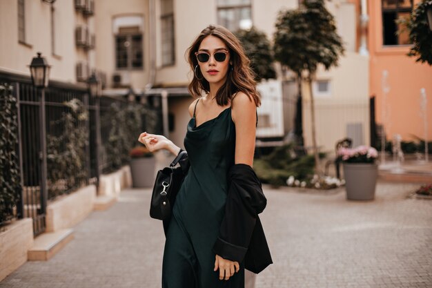 Modna blada brunetka w długiej zielonej sukience, czarnej kurtce i okularach przeciwsłonecznych, stojąca na ulicy w ciągu dnia przy ścianie jasnego budynku miejskiego