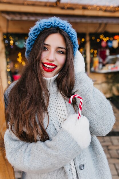 Modna brunetka kobieta w białych rękawiczkach, ciesząc się zimą. Zewnątrz portret wyrafinowanej długowłosej modelki w stylowym niebieskim kapeluszu pozuje z przyjemnością w zimny poranek, trzymając świąteczne cukierki.