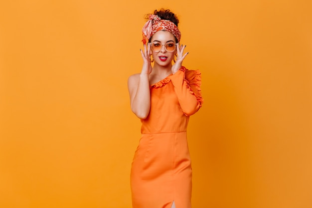Modna ciemnowłosa dama w afrykańskiej opasce, okularach przeciwsłonecznych i eleganckiej sukience zalotnie patrzy w kamerę w pomarańczowej przestrzeni.