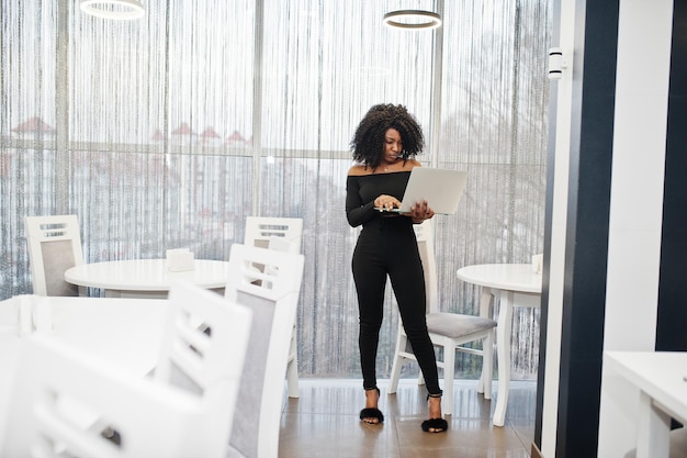 Modna młoda piękna afroamerykańska kobieta biznesu z fryzurą afro w eleganckim czarnym stojaku z laptopem pod ręką