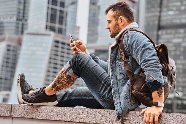 Modnie ubrany, wytatuowany mężczyzna hipster z plecakiem za pomocą smartfona siedzący przed wieżowcami w mieście Mosków w pochmurny poranek.